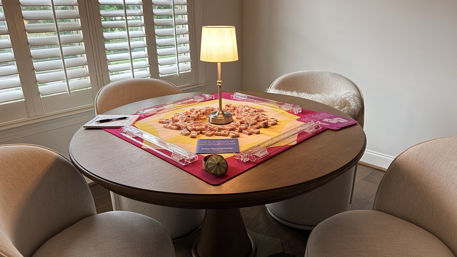Round wood table set for mahjong with blush tiles on a hot-pink and citron mat, acrylic racks, and a small lamp glowing at the center, surrounded by four cream chairs