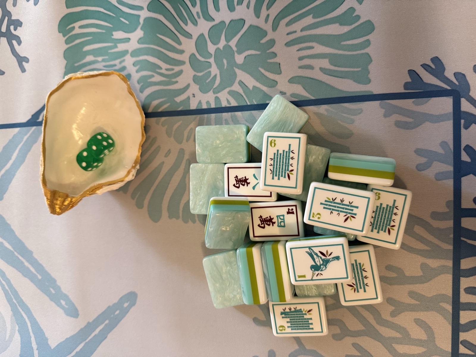 Seafoam, aqua, olive, and white mahjong tiles piled on a blue coral patterned mat, with a gold-rimmed oyster shell holding two green dice
