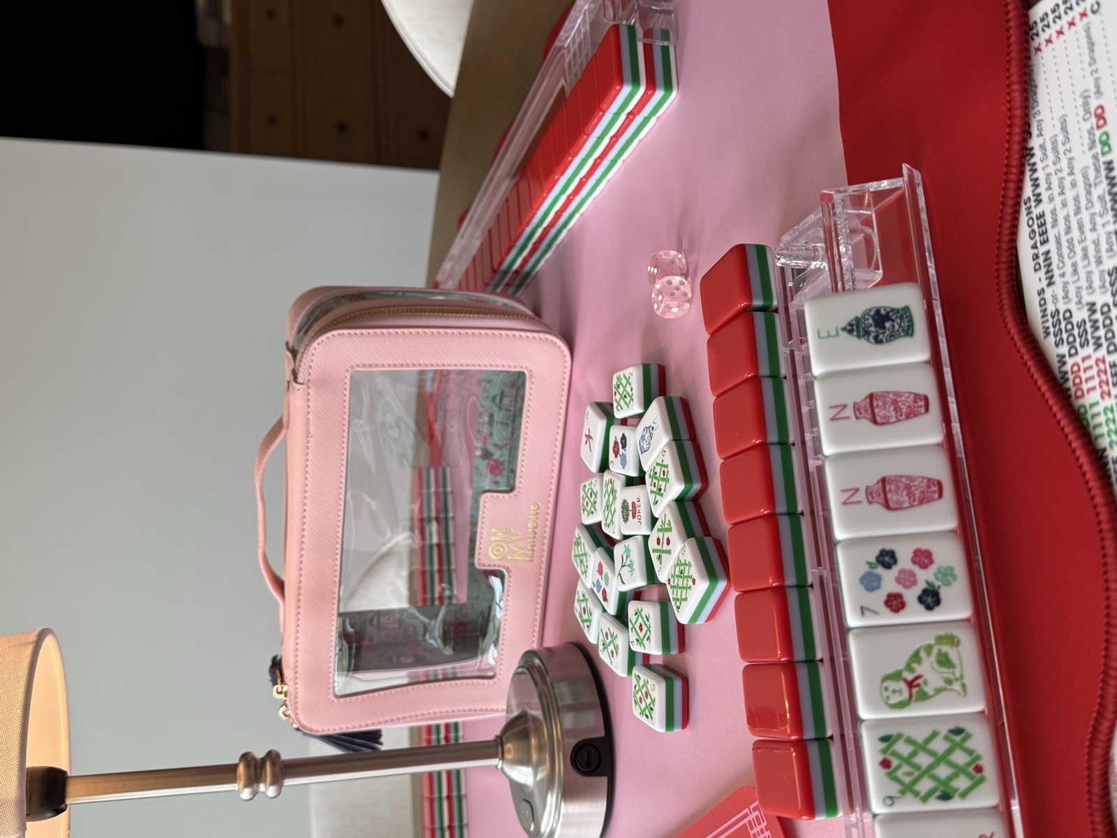 Pink Oh My Mahjong zip pouch beside red-and-green Nantucket tiles and a rack holding a Staffordshire dog, floral, and ginger jar tiles on a pink mat