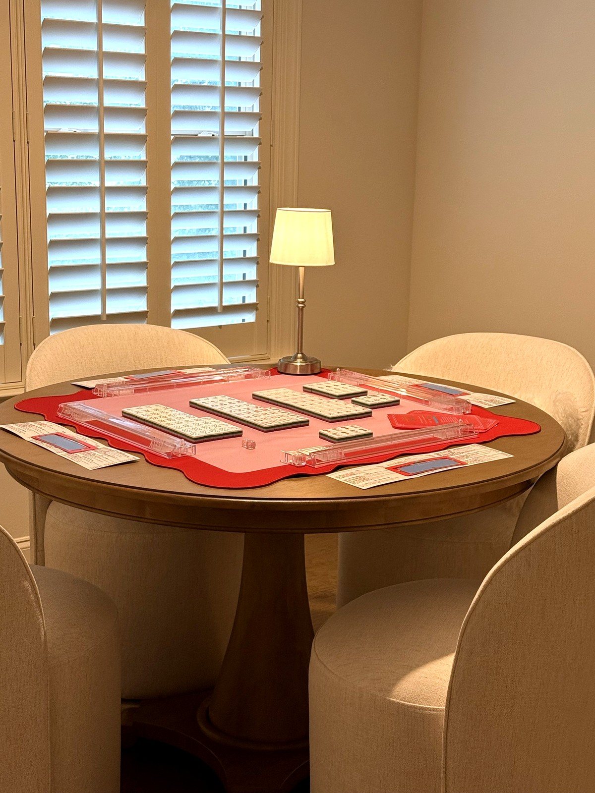 Round wood table set for mahjong with a pink-and-red scalloped mat, clear lucite racks and pushers, red-and-green tiles, and a small lamp glowing by a shuttered window
