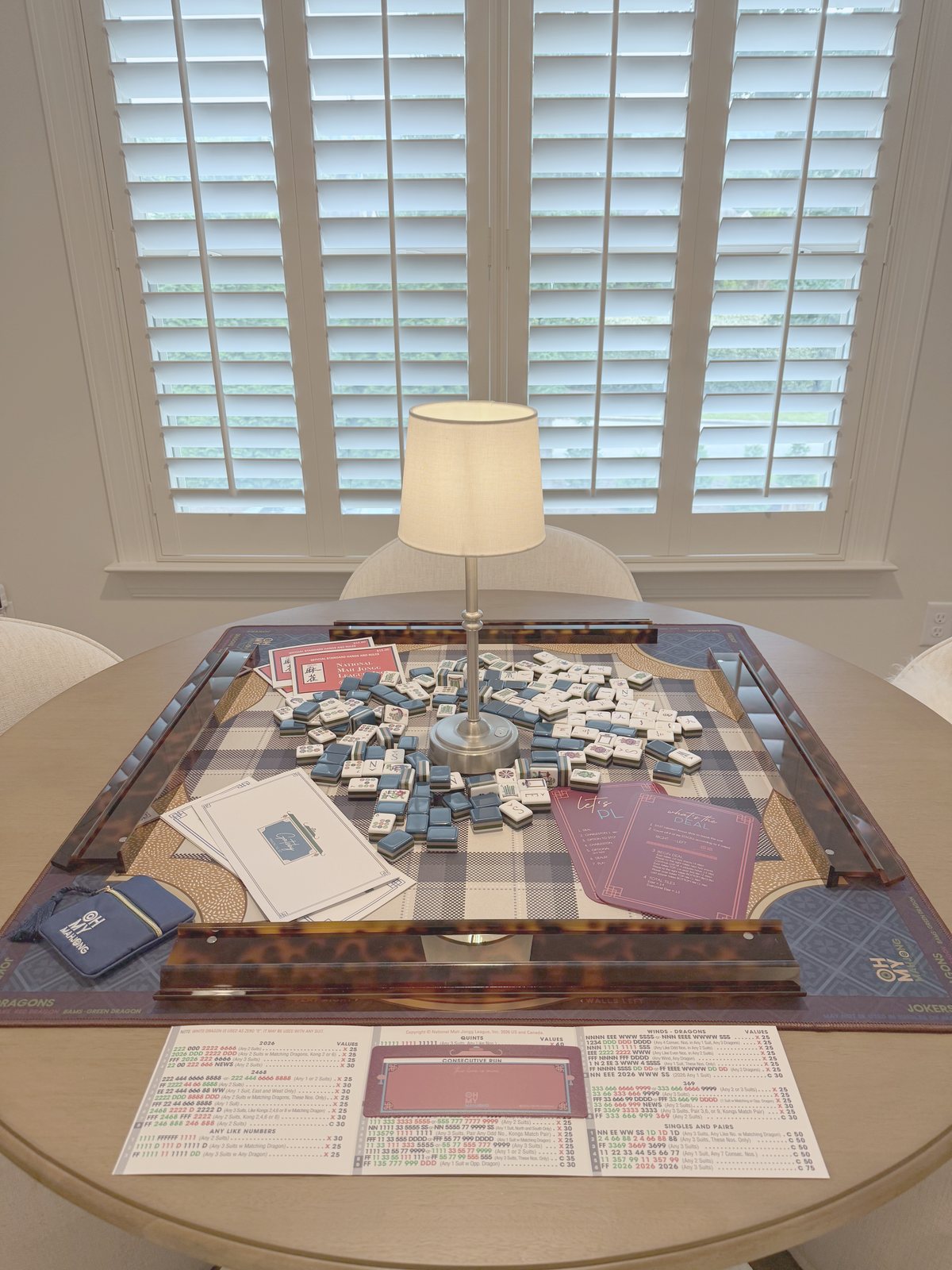 Round wood table set for mahjong with a navy buffalo-check mat, tortoiseshell racks, navy tiles, and a small lamp glowing at the center