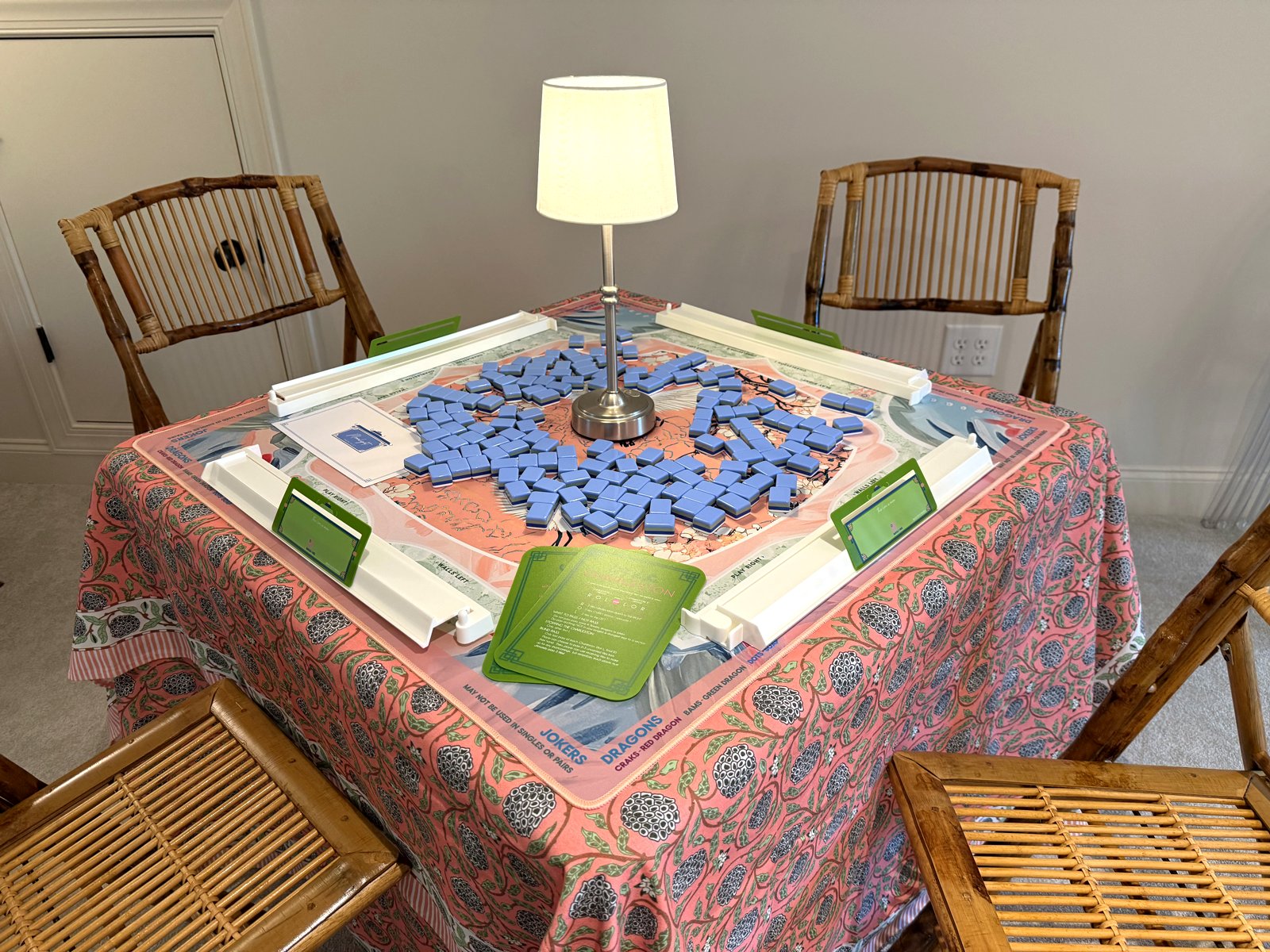 Square mahjong table with a coral Oh My Mahjong Heron mat, cornflower-blue Moonlight tiles, white acrylic racks and pushers, green Charleston cards, and a small lamp, surrounded by bamboo folding chairs