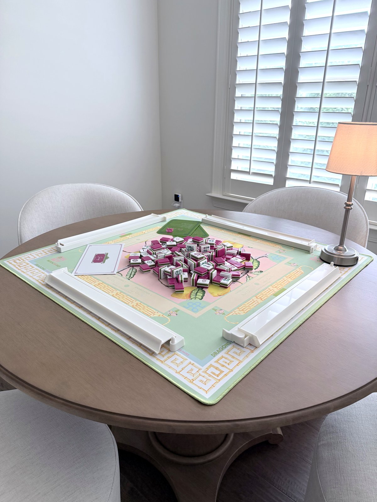 Round wood pedestal table set for mahjong with Oh My Mahjong's mint-and-citron Citrus Songbird mat, magenta-and-green Dandy tiles, white acrylic racks and pushers, and a lamp by plantation shutters